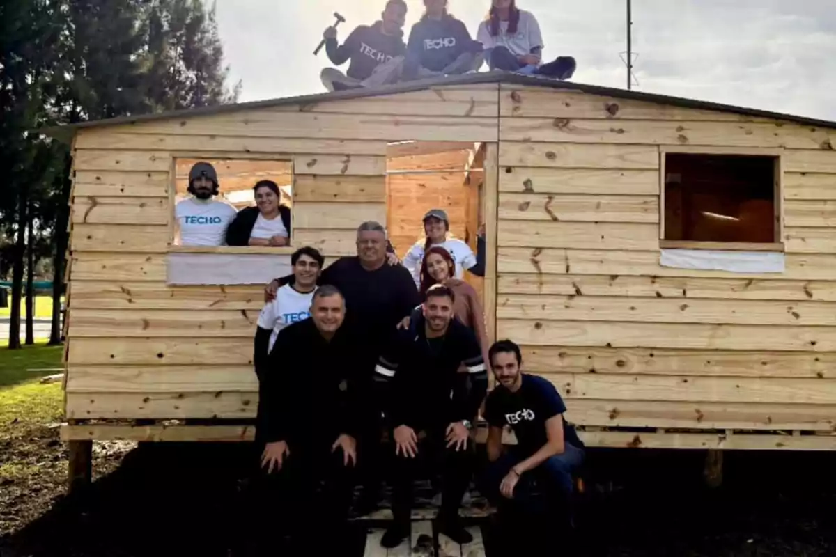 Grupo de personas sonrientes posando frente a una casa de madera recién construida, algunas de ellas están sentadas en el techo y otras en la entrada, en un entorno al aire libre con árboles al fondo