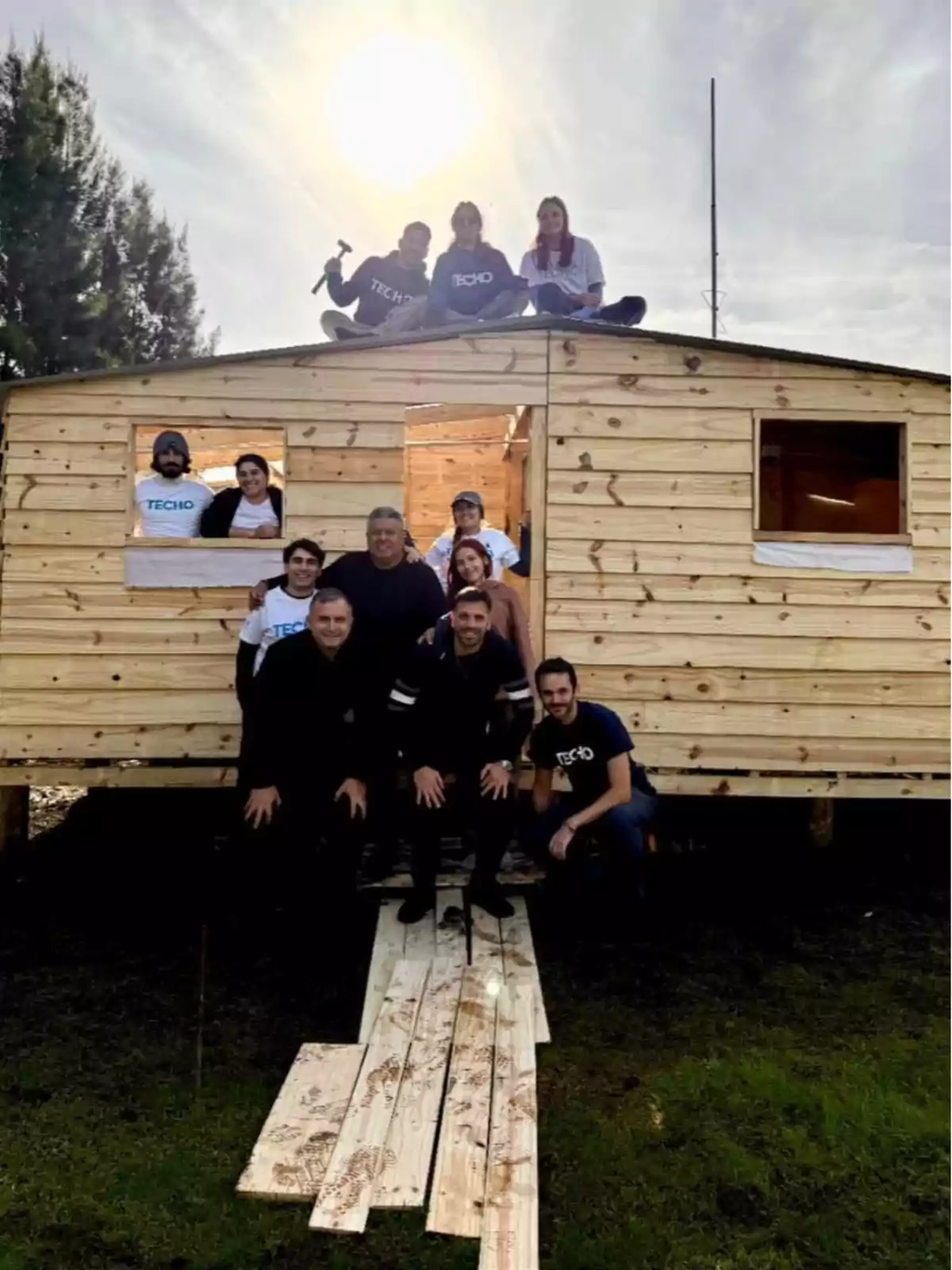 Grupo de personas posando frente a una casa de madera recién construida, algunas están sentadas en el techo y otras de pie en la entrada, con el sol brillando en el fondo