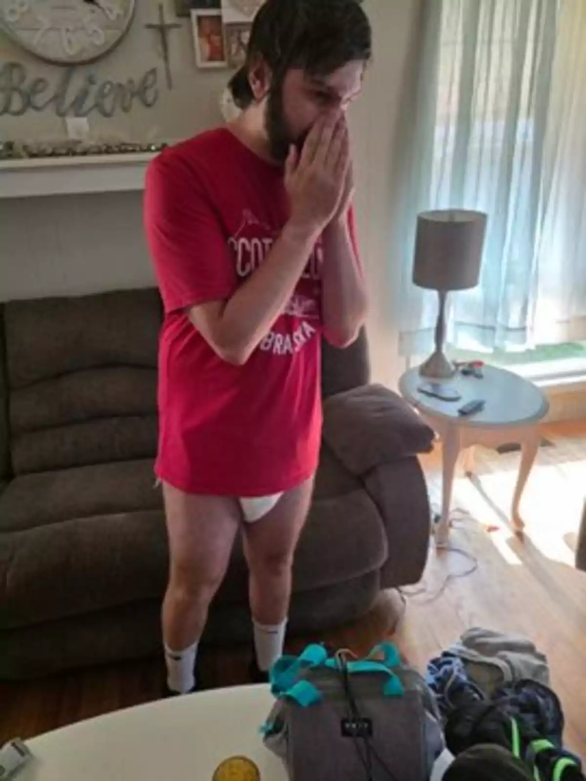Joven de pie en una sala con expresión de sorpresa, usando camiseta roja, calcetas blancas y ropa interior, con las manos cubriéndose la boca. Joven de pie en una sala con expresión de sorpresa, usando camiseta roja, calcetas blancas y ropa interior, con las manos cubriéndose la boca.