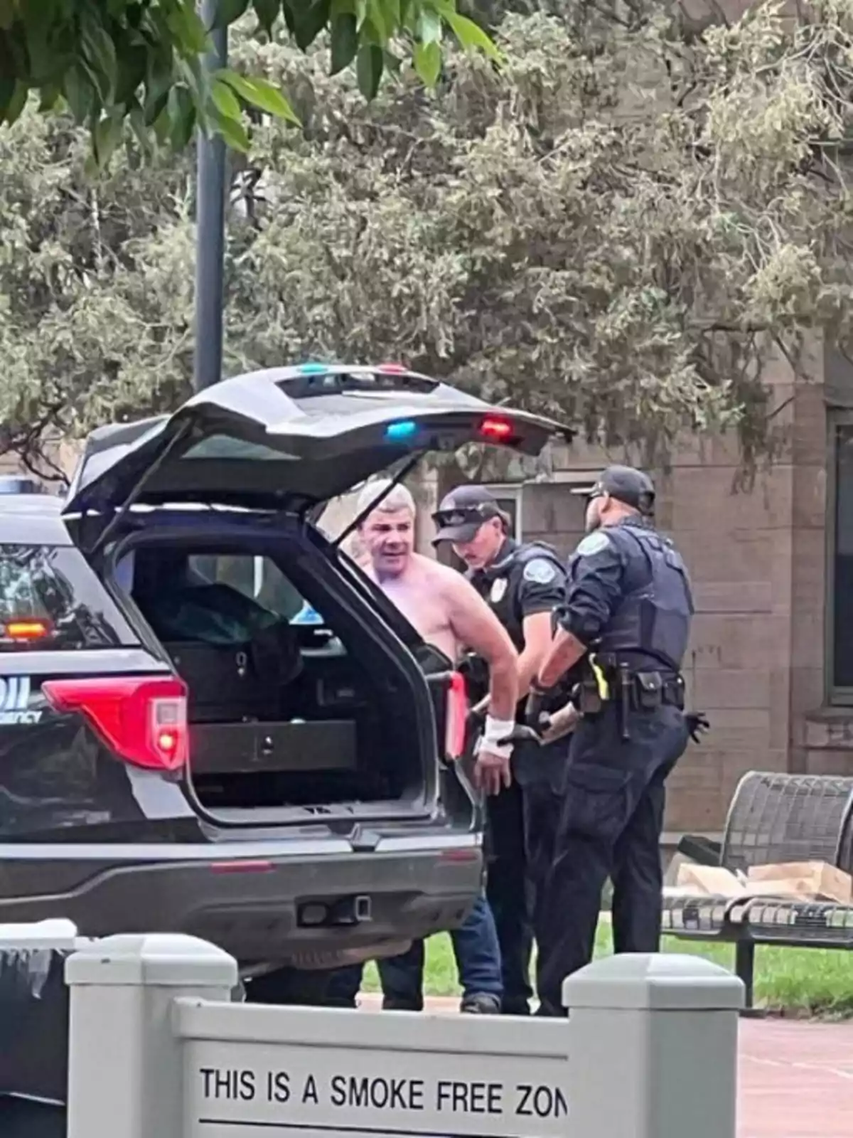 Un hombre sin camisa está siendo arrestado por dos policías junto a una patrulla con la cajuela abierta en una zona donde no se permite fumar. Un hombre sin camisa está siendo arrestado por dos policías junto a una patrulla con la cajuela abierta en una zona donde no se permite fumar.