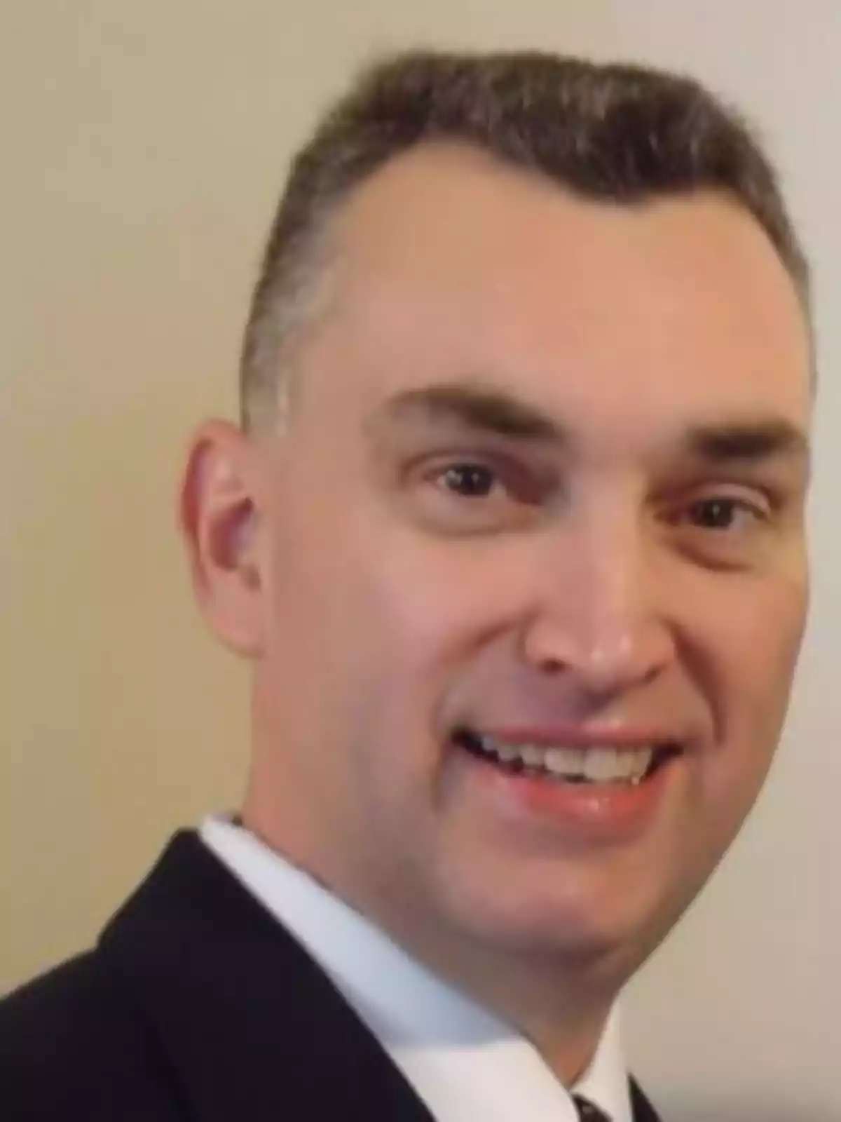 Short dark-haired man wearing a formal suit and smiling in front of a light-colored wall Short dark-haired man wearing a formal suit and smiling in front of a light-colored wall