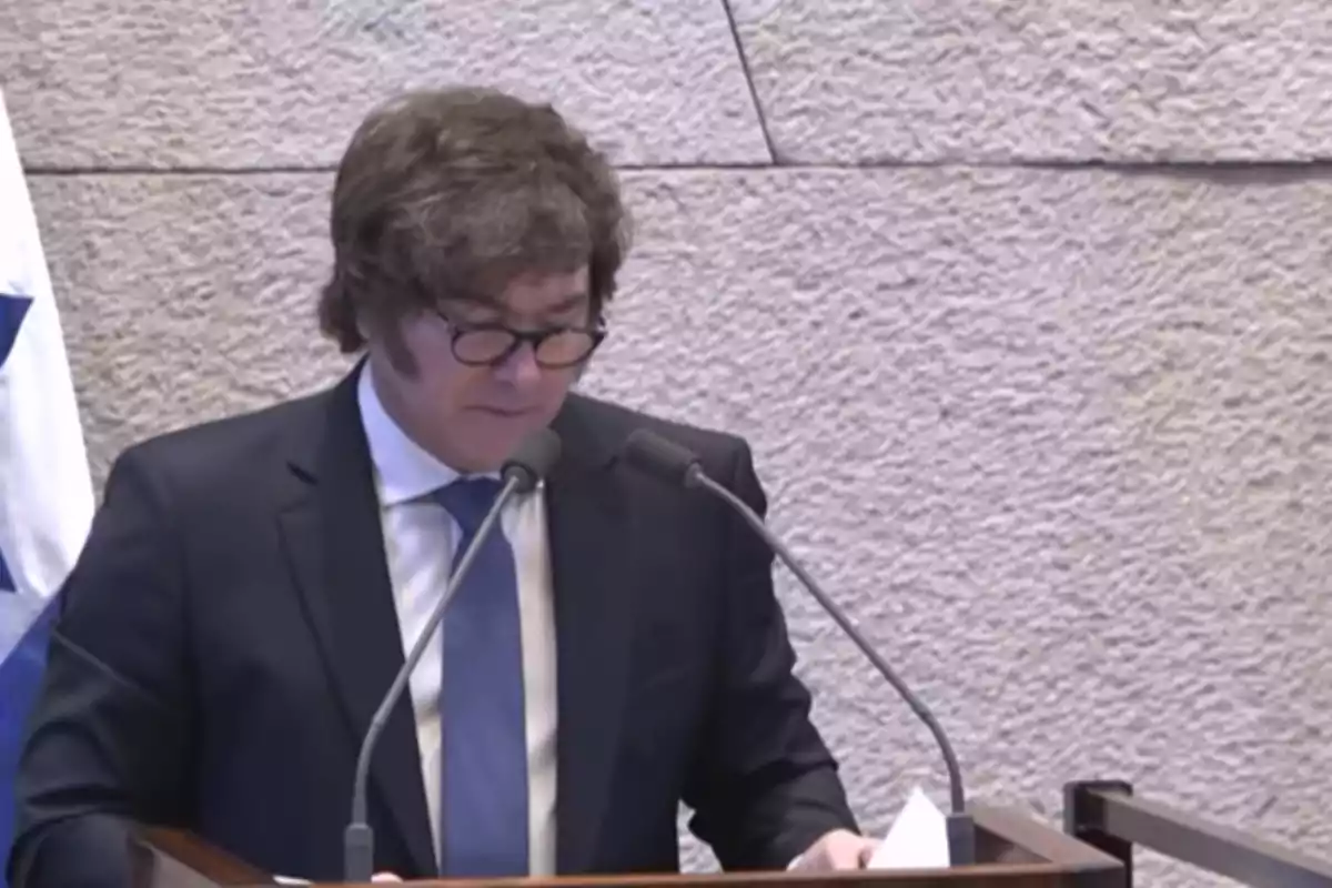 Hombre con gafas y traje oscuro hablando en un podio con dos micrófonos frente a una pared de piedra y una bandera blanca con azul a la izquierda Hombre con gafas y traje oscuro hablando en un podio con dos micrófonos frente a una pared de piedra y una bandera blanca con azul a la izquierda