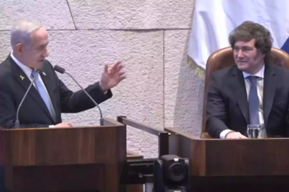 Dos hombres con traje conversan en un recinto oficial uno de pie hablando en un podio y el otro sentado escuchando Dos hombres con traje conversan en un recinto oficial uno de pie hablando en un podio y el otro sentado escuchando
