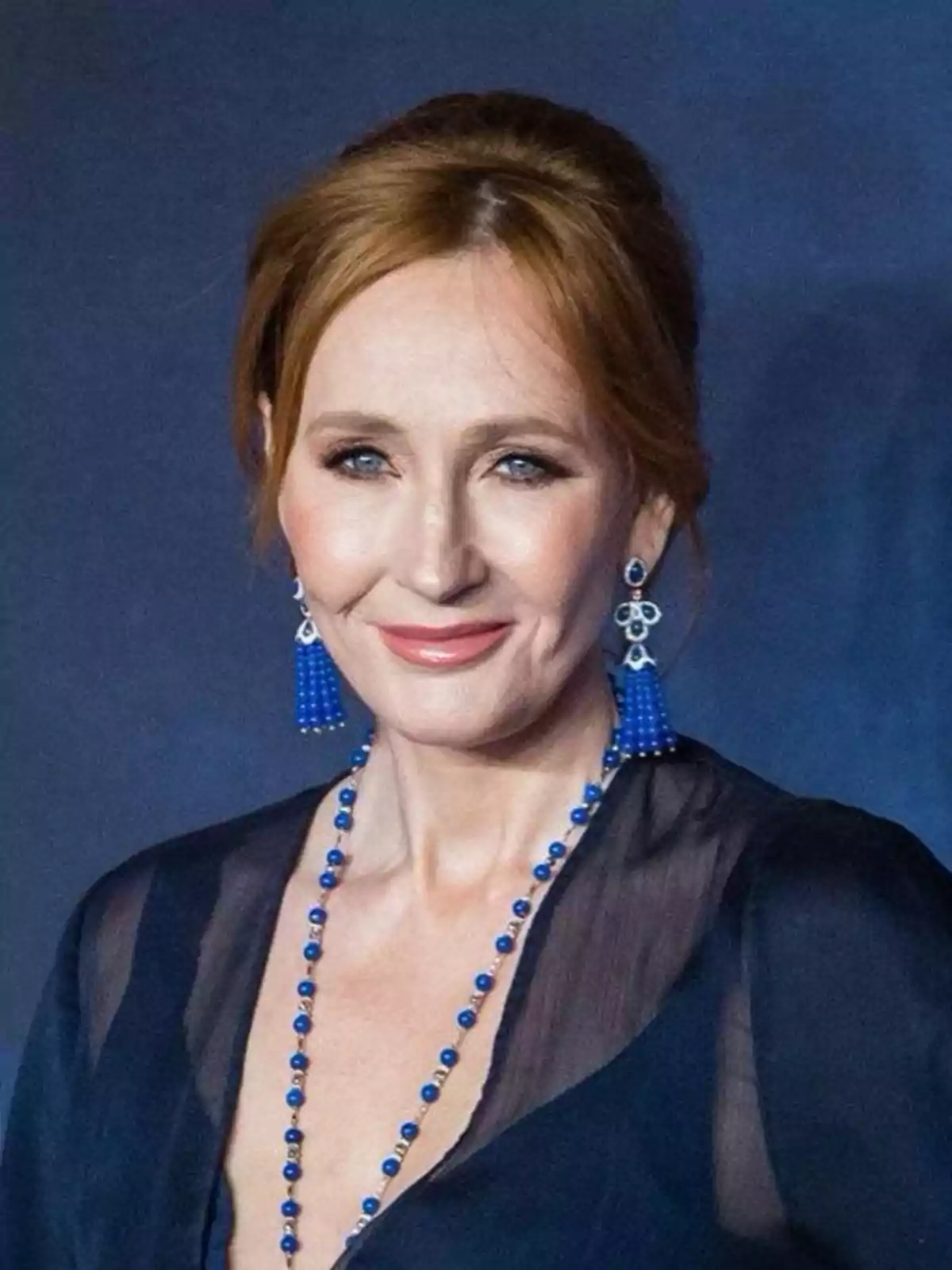 Red-haired woman with her hair up, wearing blue earrings and necklace, elegantly dressed and smiling in front of a dark background Red-haired woman with her hair up, wearing blue earrings and necklace, elegantly dressed and smiling in front of a dark background