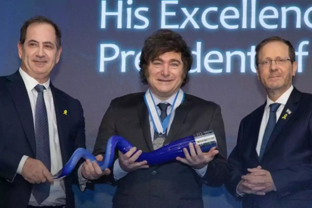 Tres hombres con traje posan sonrientes en un evento, el hombre del centro sostiene un objeto azul y lleva una medalla en el cuello