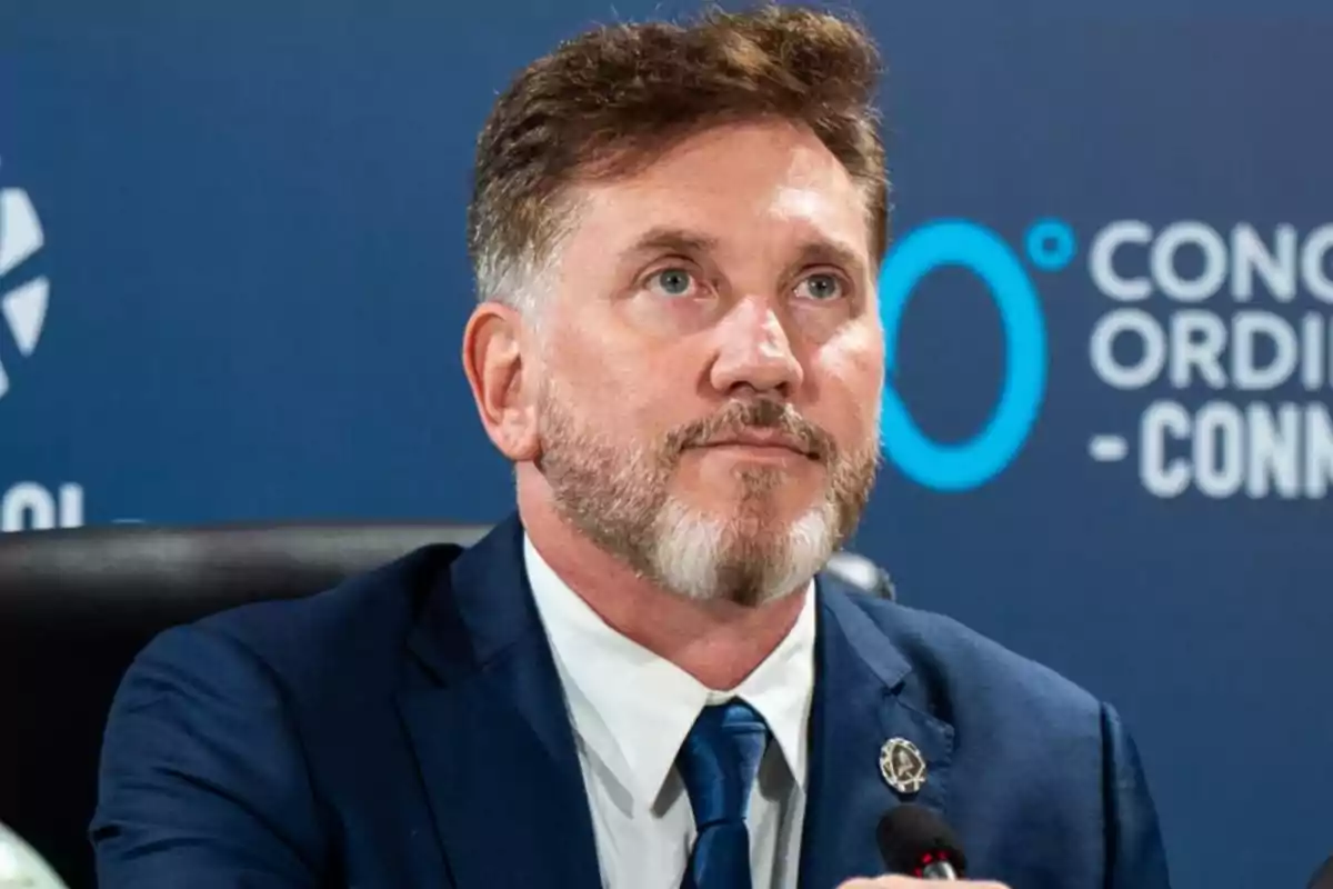 Hombre de barba y cabello corto con traje azul sentado frente a un micrófono en un evento formal con fondo azul y letras blancas