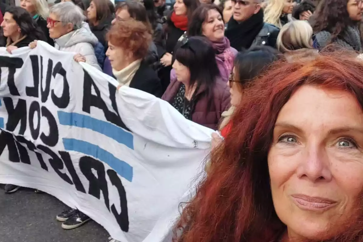 Mujer de cabello rojizo toma una selfie en una manifestación donde varias personas sostienen una pancarta blanca con letras negras y dos franjas celestes Mujer de cabello rojizo toma una selfie en una manifestación donde varias personas sostienen una pancarta blanca con letras negras y dos franjas celestes