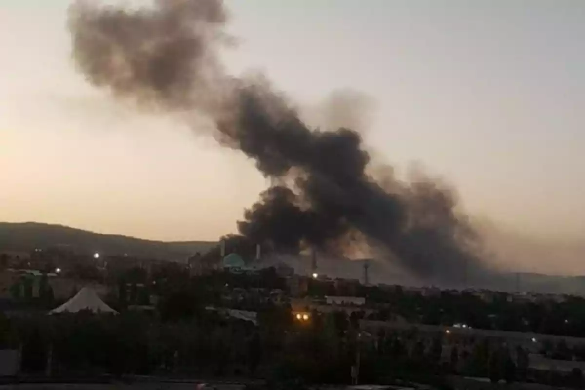 Una gran columna de humo negro se eleva sobre una ciudad al atardecer, indicando un posible incendio o explosión en la zona urbana.