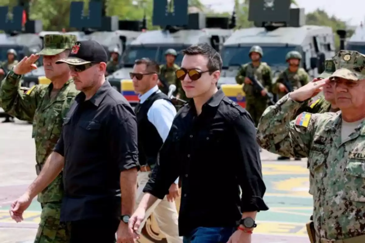 A group of people, some in military uniform, walk past armored vehicles while two of them salute.