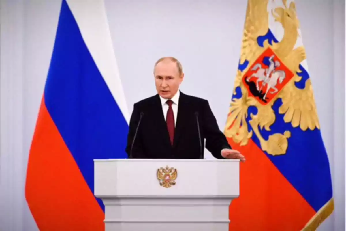 A man in a dark suit gives a speech at a podium in front of a Russian flag and a banner with a golden eagle.