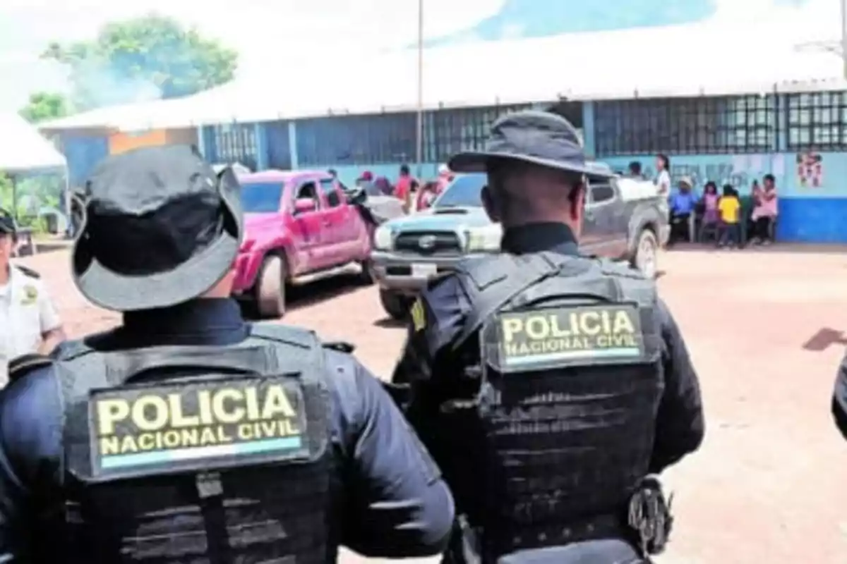 Two National Civil Police officers with their backs turned observe a scene with parked vehicles and people gathered in front of a building.