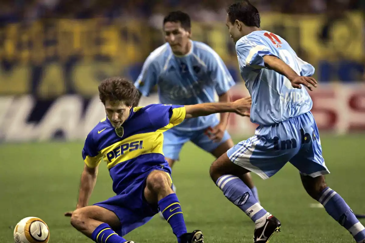 Soccer players compete for the ball in a match, one of them wearing a blue and yellow uniform while the other two are dressed in light blue. Soccer players compete for the ball in a match, one of them wearing a blue and yellow uniform while the other two are dressed in light blue.
