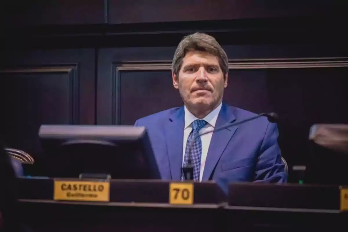 Un hombre con traje azul y corbata está sentado en un escritorio oscuro con una placa que dice "Castello Guillermo" y el número 70.