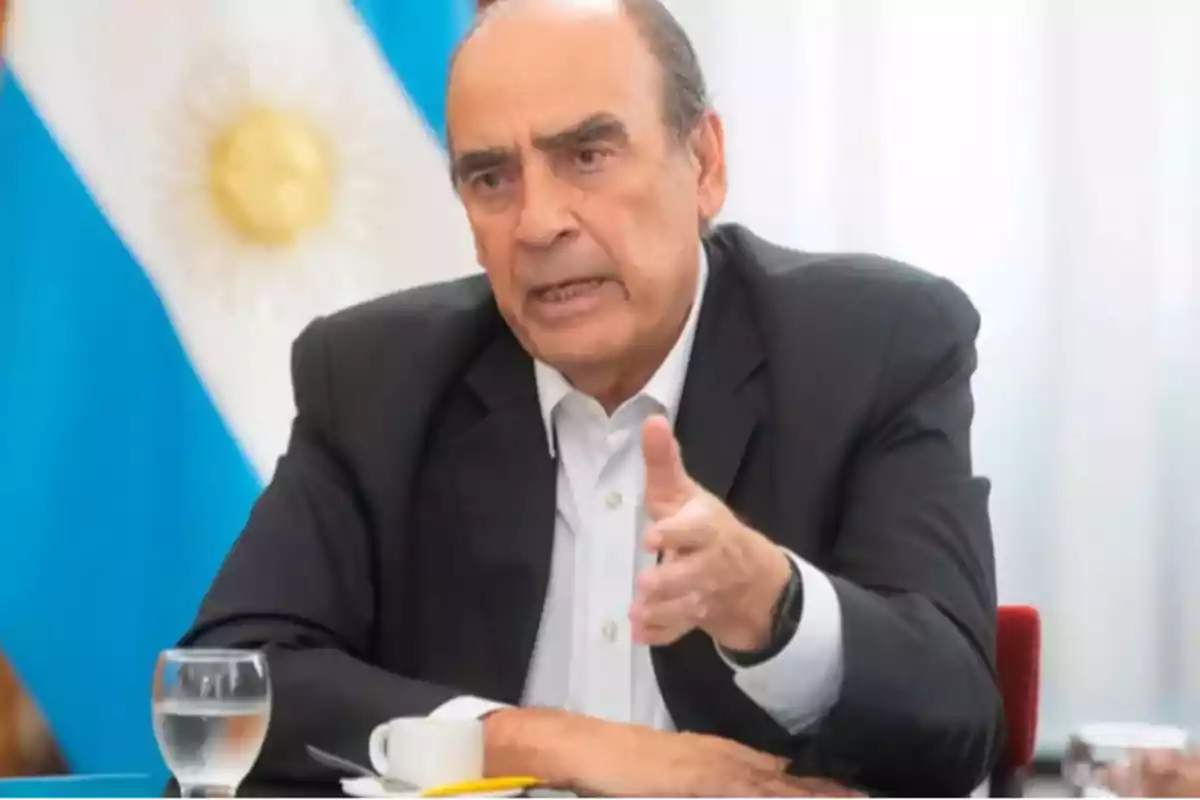 Un hombre de traje oscuro gesticula mientras habla, con una bandera argentina de fondo.