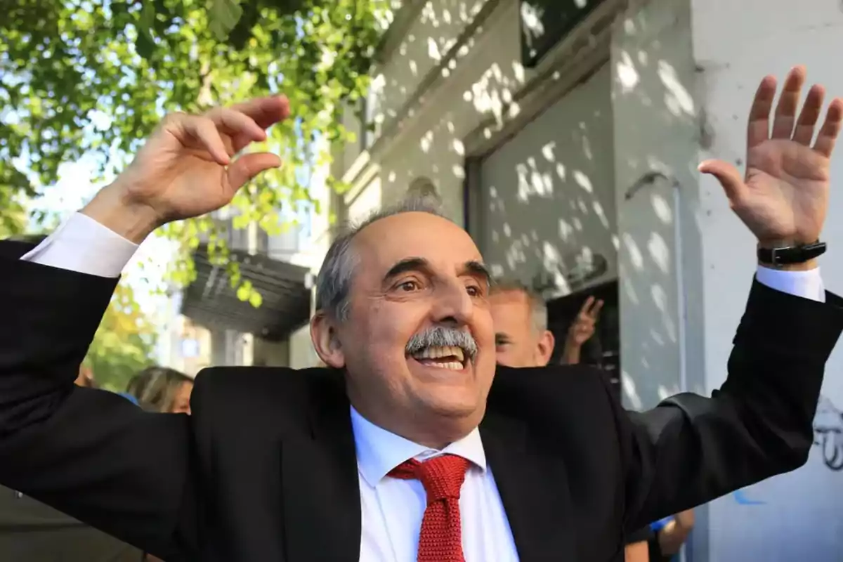 Un hombre sonriente con traje y corbata roja levanta los brazos en un gesto de celebración al aire libre.