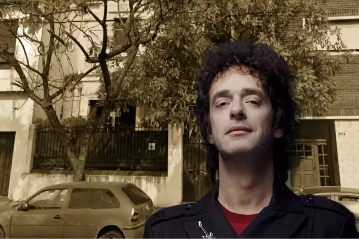 Hombre de cabello rizado y expresión seria posando frente a una casa con árboles y un auto estacionado
