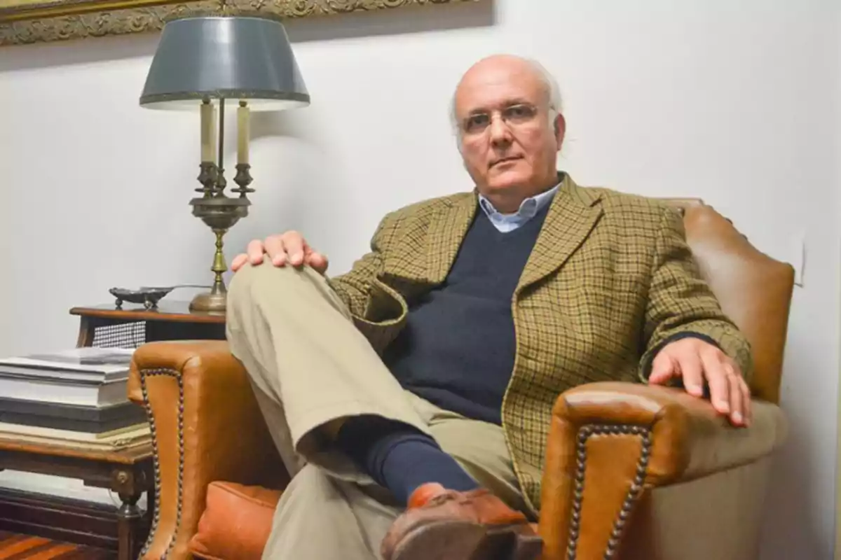 Hombre mayor sentado en un sillón de cuero marrón con las piernas cruzadas, vistiendo saco a cuadros, suéter oscuro y pantalón claro, en una sala con lámpara y libros sobre una mesa auxiliar Hombre mayor sentado en un sillón de cuero marrón con las piernas cruzadas, vistiendo saco a cuadros, suéter oscuro y pantalón claro, en una sala con lámpara y libros sobre una mesa auxiliar