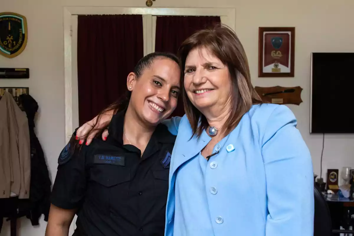 Dos mujeres sonrientes posan juntas en una habitación con decoraciones en la pared. Una lleva uniforme y la otra un traje azul.