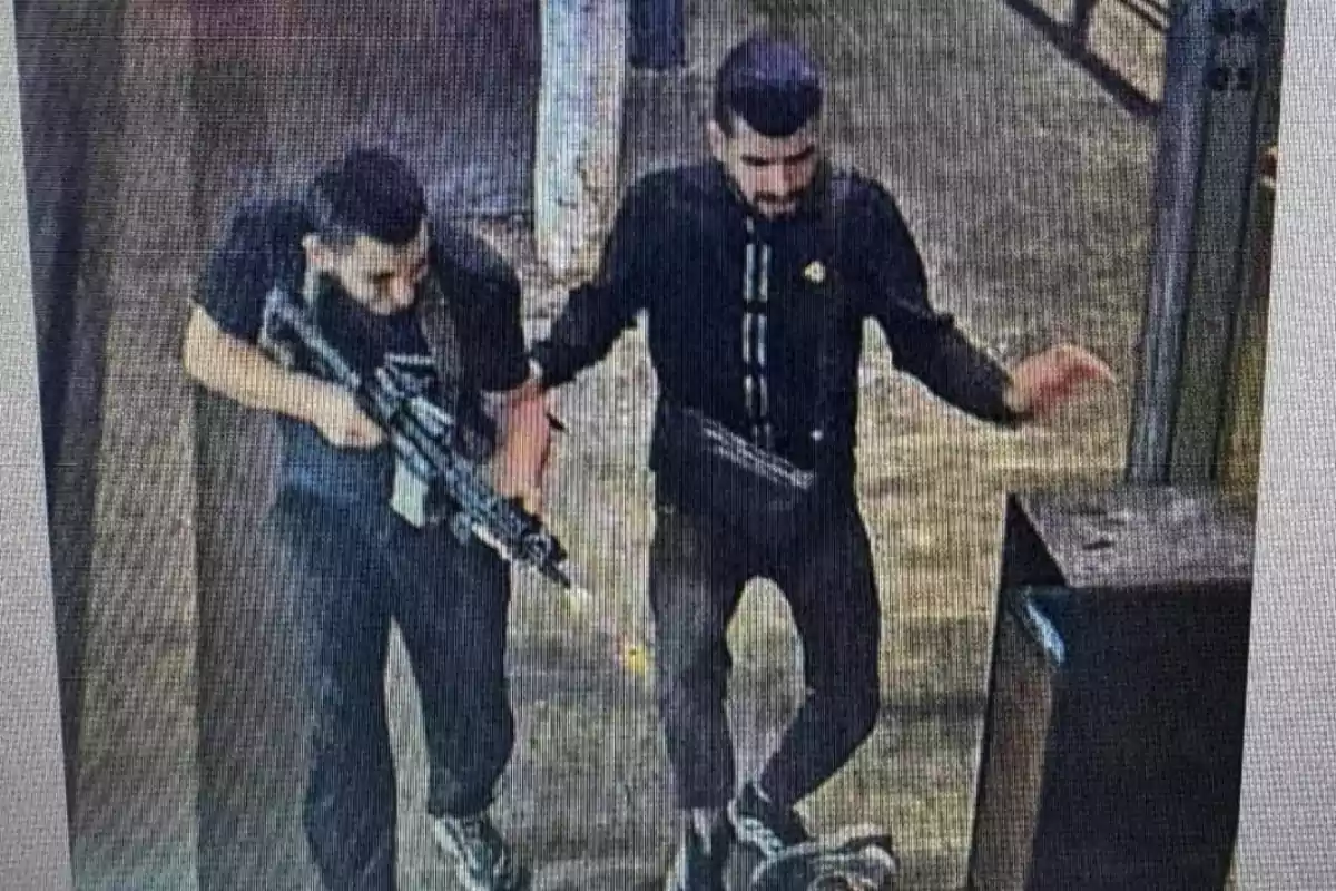 Dos hombres vestidos de negro, uno de ellos portando un arma larga, caminando juntos en un entorno urbano.