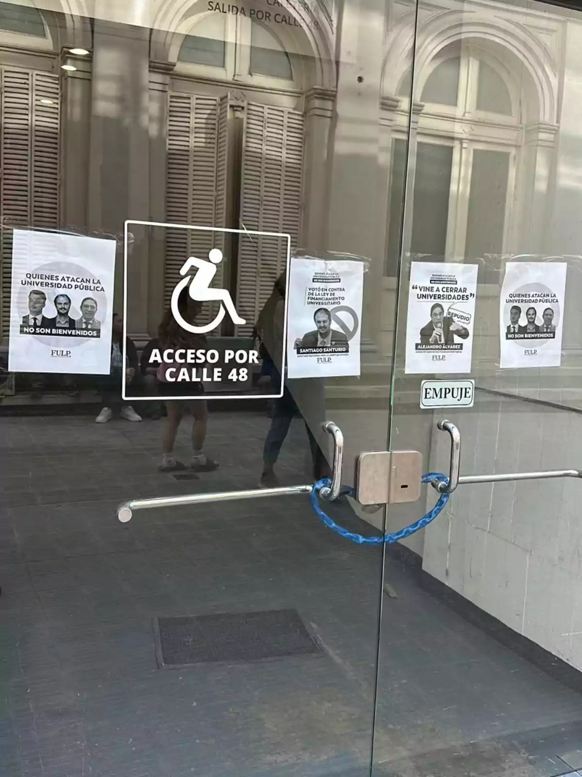 Puerta de vidrio con carteles de protesta y un símbolo de acceso para personas con discapacidad, asegurada con una cadena azul.