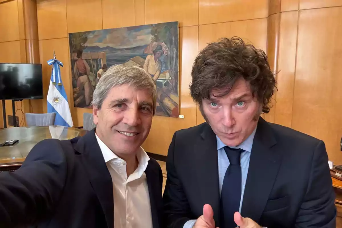 Two men in suits pose for a selfie in an office with an Argentine flag and a painting on the wall.