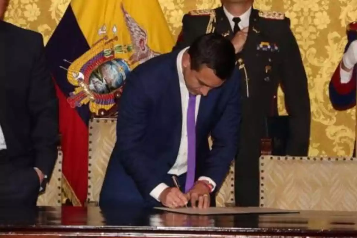 A man signing a document at an official ceremony with the flag of Ecuador in the background.