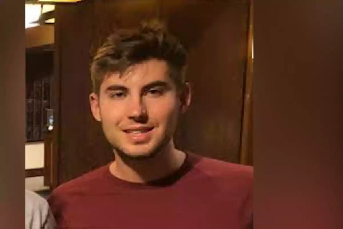 Un hombre joven con cabello castaño y barba incipiente, vistiendo una camiseta marrón, está de pie frente a un fondo de madera.