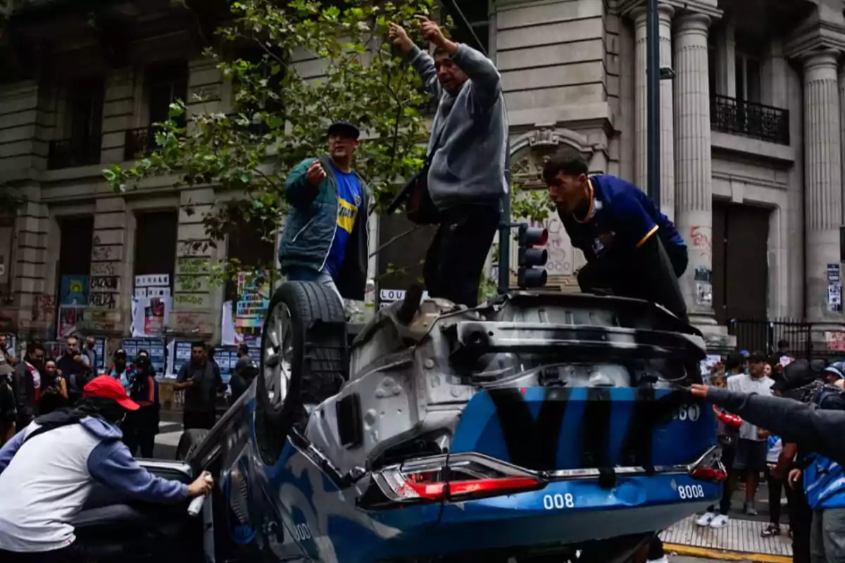 A group of people is on top of an overturned car in an urban street, surrounded by old buildings and graffiti on the walls, while some watch the scene.