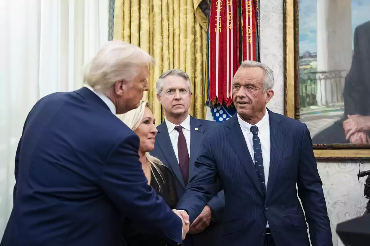 Four people dressed in suits greet each other and talk in an elegant room with golden curtains and a painting on the wall.