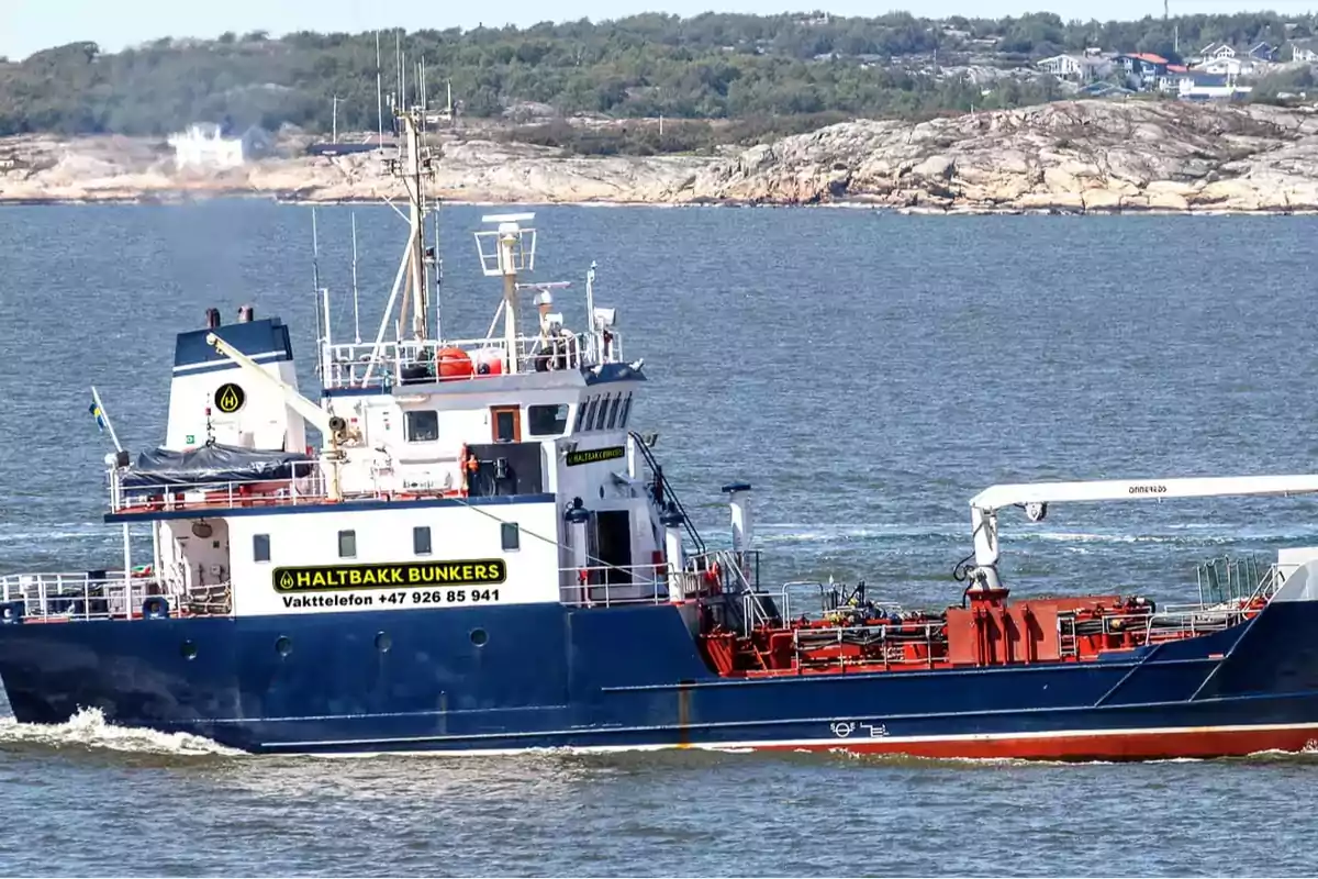 A blue and white cargo ship sails near a rocky coast with vegetation. A blue and white cargo ship sails near a rocky coast with vegetation.
