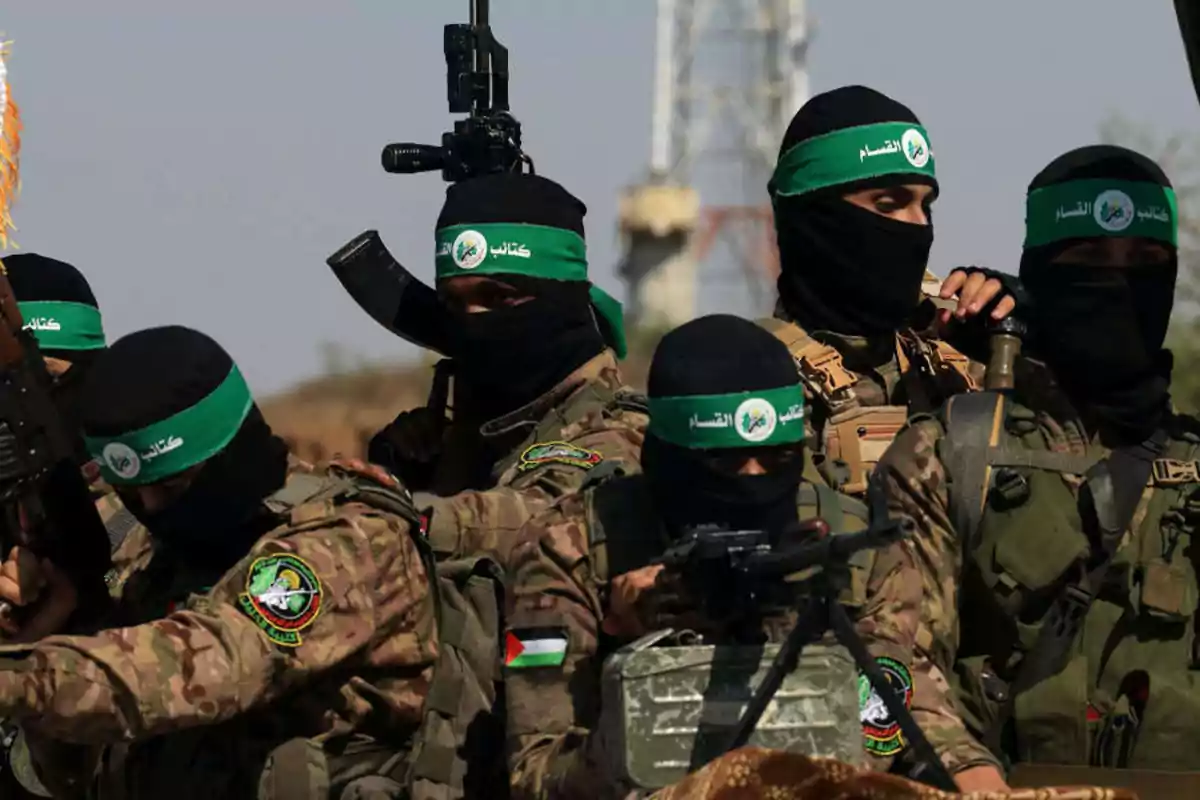 Several armed men wearing camouflage uniforms and black balaclavas with green bands are gathered outdoors. Several armed men wearing camouflage uniforms and black balaclavas with green bands are gathered outdoors.