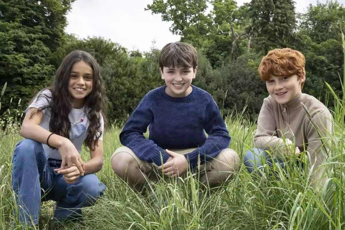 Tres niños sonríen mientras están agachados en un campo de pasto alto con árboles al fondo