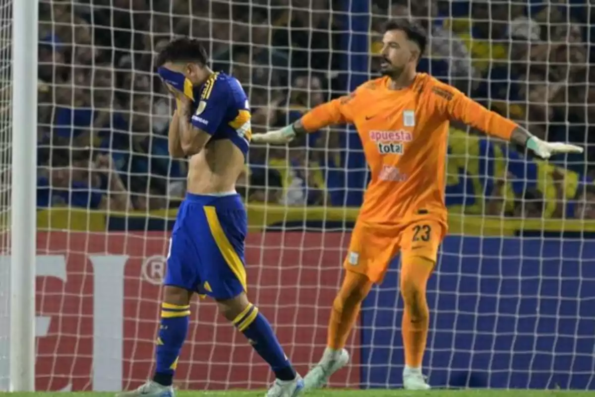 Un jugador de fútbol se cubre el rostro con la camiseta mientras un portero con uniforme naranja extiende los brazos en el área de juego. Un jugador de fútbol se cubre el rostro con la camiseta mientras un portero con uniforme naranja extiende los brazos en el área de juego.