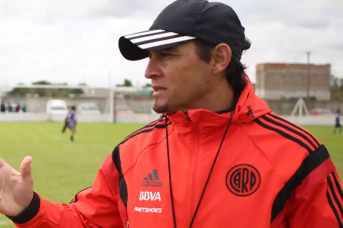 A man with a red jacket and a black cap on a soccer field.