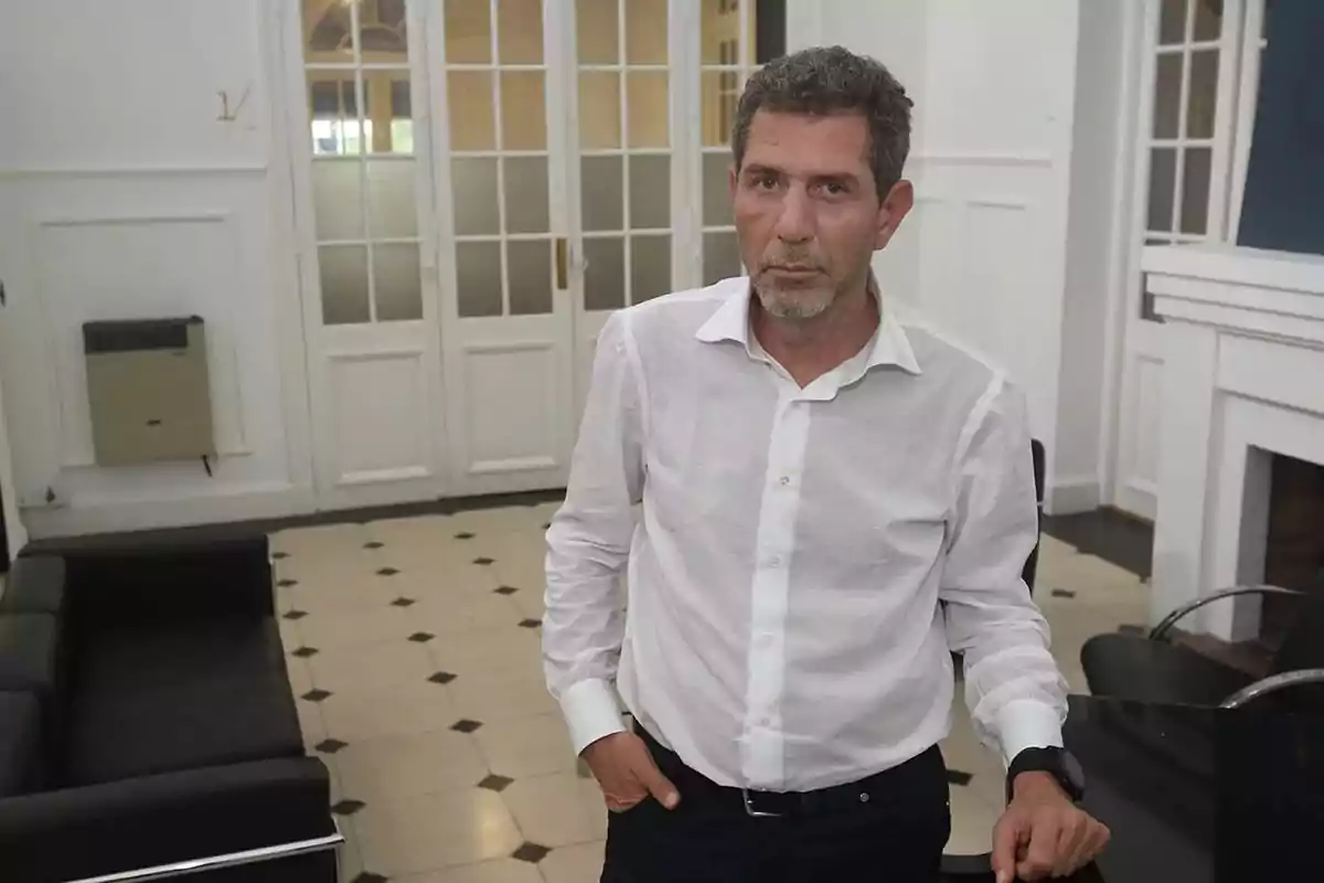 Un hombre de pie en una sala con puertas de vidrio al fondo, vistiendo una camisa blanca. Un hombre de pie en una sala con puertas de vidrio al fondo, vistiendo una camisa blanca.