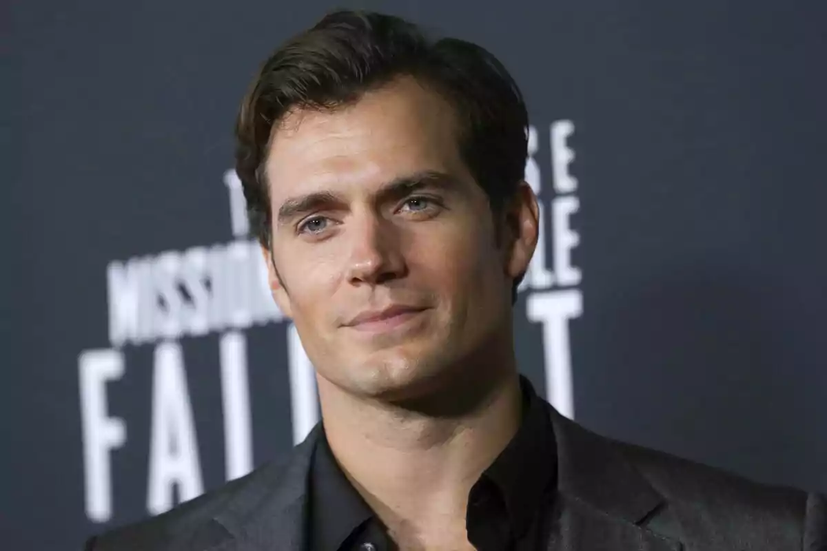Hombre de cabello oscuro con traje gris posando frente a un fondo con letras blancas desenfocadas Hombre de cabello oscuro con traje gris posando frente a un fondo con letras blancas desenfocadas
