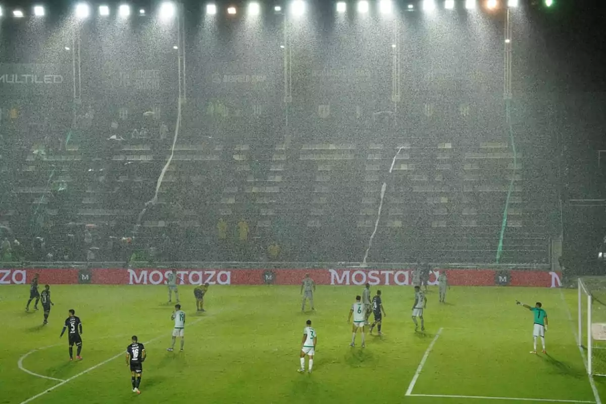 Soccer players on a field under heavy rain during a night game with bright lights illuminating the stadium and partially empty stands.