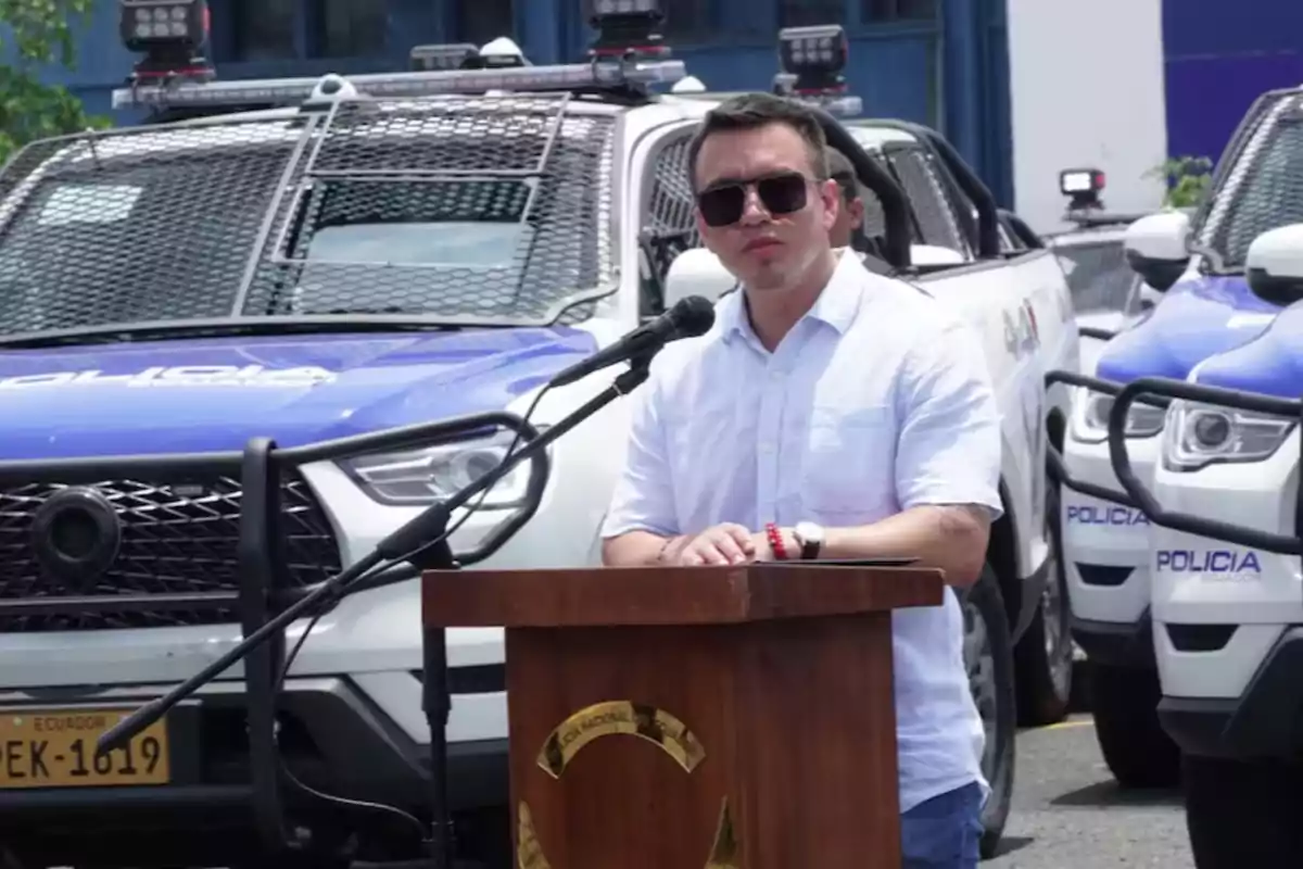 A man with sunglasses speaks at a podium in front of police vehicles.