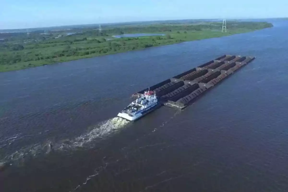 Una embarcación empuja varias barcazas cargadas navegando por un río amplio con vegetación en las orillas bajo un cielo despejado. Una embarcación empuja varias barcazas cargadas navegando por un río amplio con vegetación en las orillas bajo un cielo despejado.