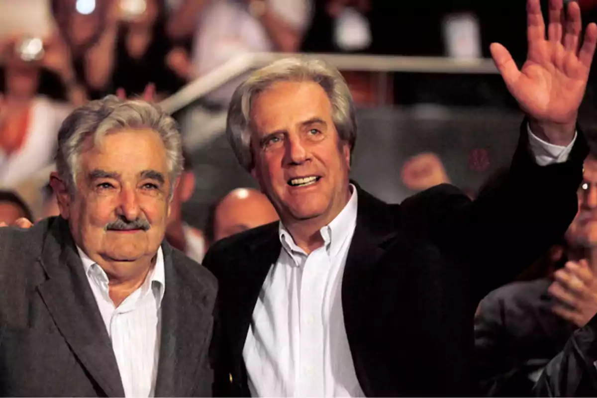 Two men standing, one of them raising his hand, at an event with an audience in the background.