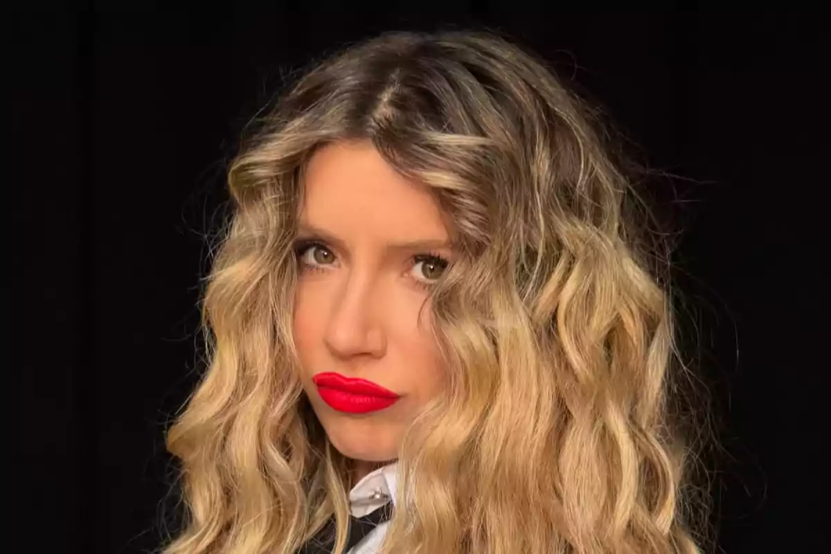 Una persona con cabello rizado rubio y labios rojos en un fondo oscuro.