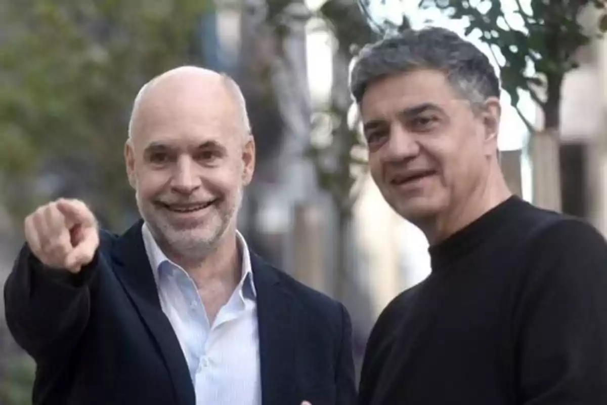 Dos hombres sonrientes en un entorno urbano, uno de ellos señalando hacia adelante.