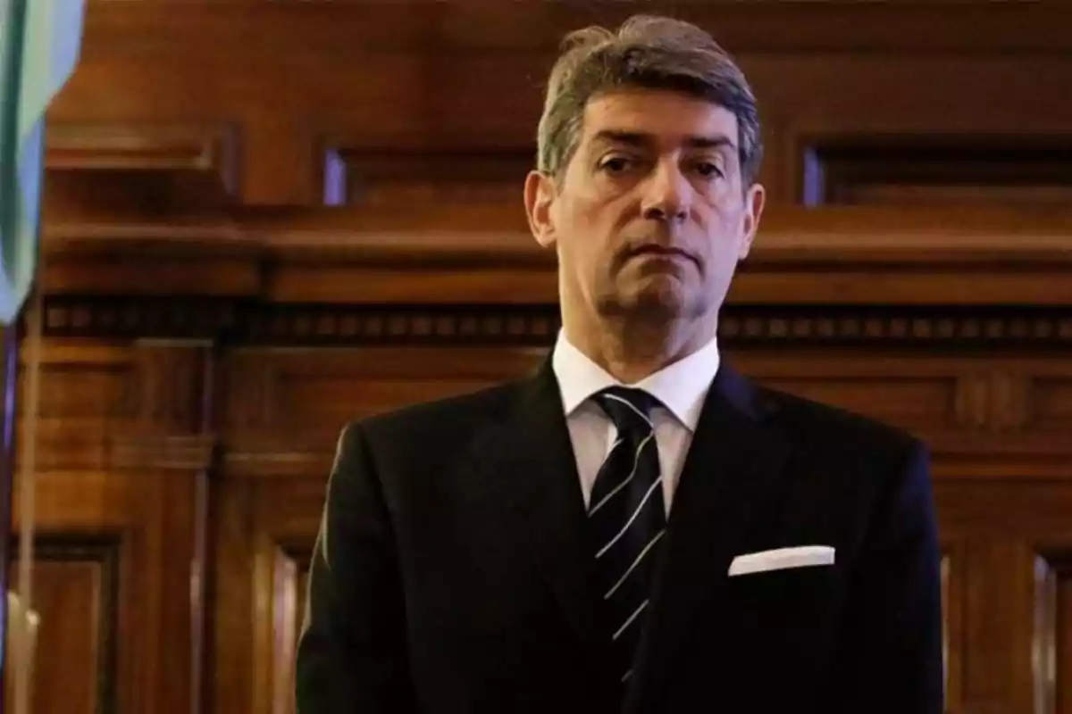 Hombre de cabello canoso vistiendo traje oscuro y corbata, de pie en un ambiente elegante con paredes de madera. Hombre de cabello canoso vistiendo traje oscuro y corbata, de pie en un ambiente elegante con paredes de madera.