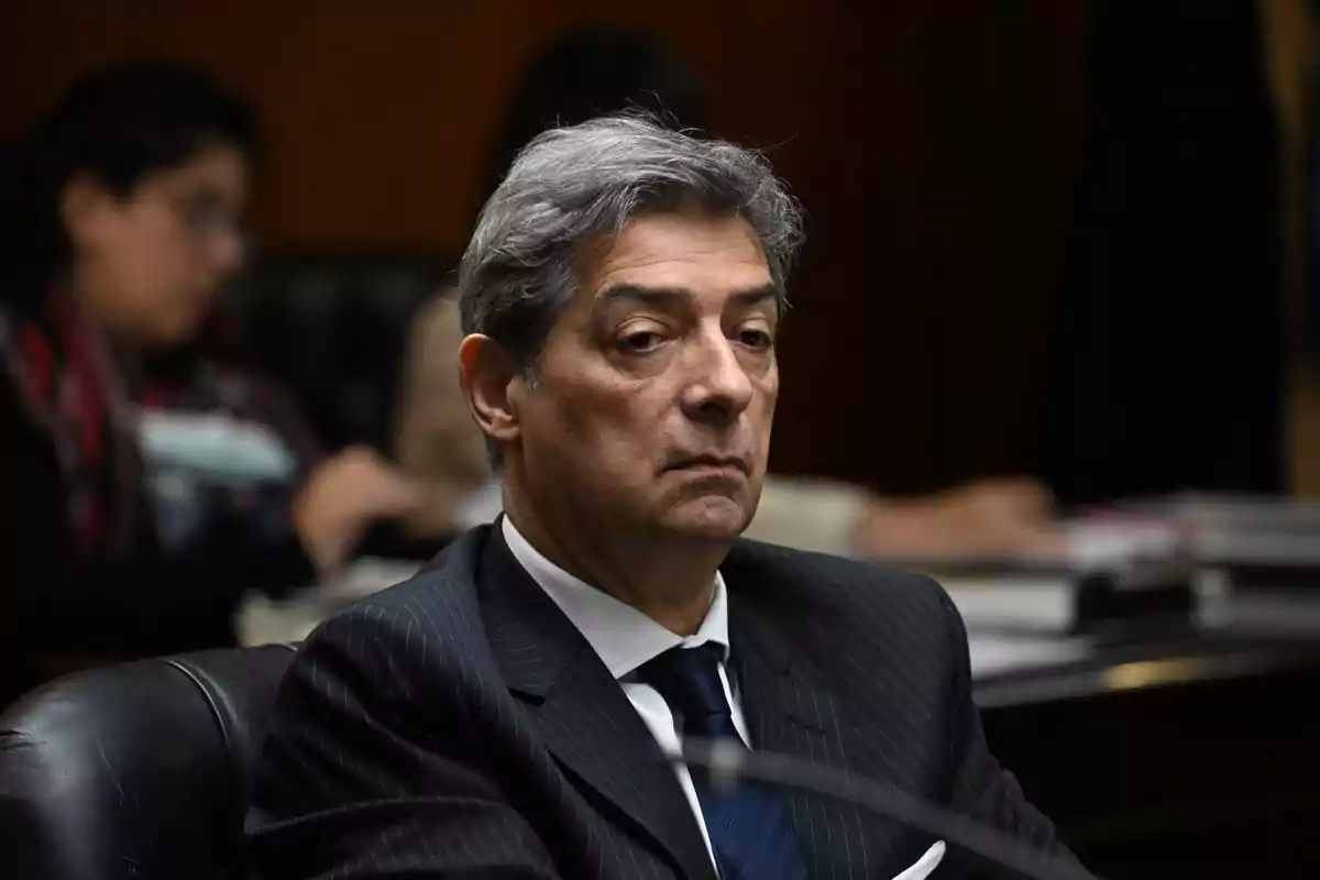 Hombre de cabello canoso con traje oscuro y corbata azul sentado en una sala de reuniones con personas y documentos al fondo desenfocados Hombre de cabello canoso con traje oscuro y corbata azul sentado en una sala de reuniones con personas y documentos al fondo desenfocados