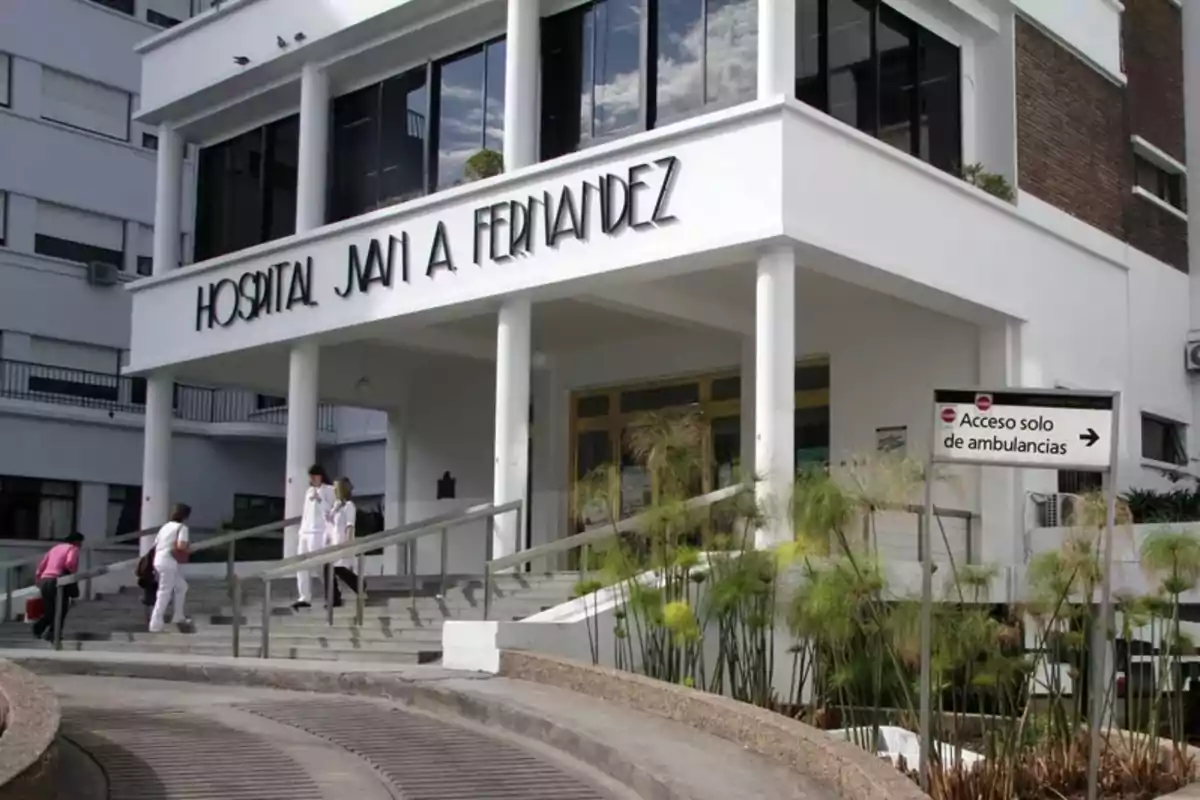 Entrada principal de un hospital con personas subiendo las escaleras y un cartel que indica acceso solo para ambulancias.