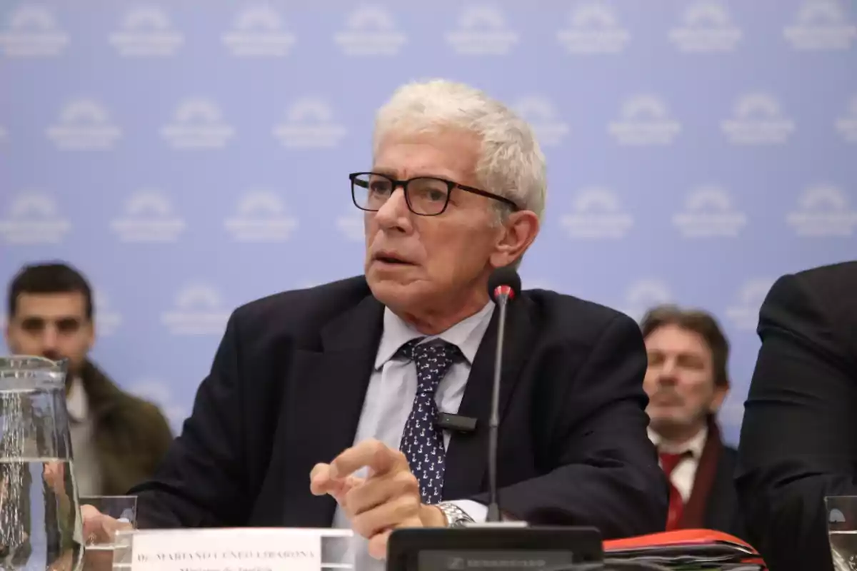 Un hombre de cabello canoso y gafas está sentado en una mesa con un micrófono frente a él, en un entorno formal con un fondo azul.