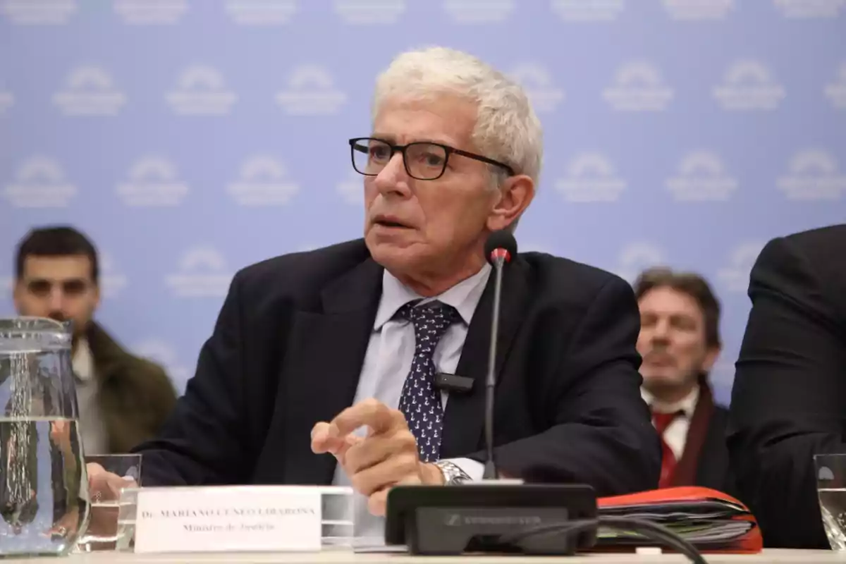 Un hombre de cabello canoso y gafas está sentado en una mesa con un micrófono frente a él, parece estar hablando o escuchando atentamente en un entorno formal, con un fondo azul que tiene un logotipo repetido.