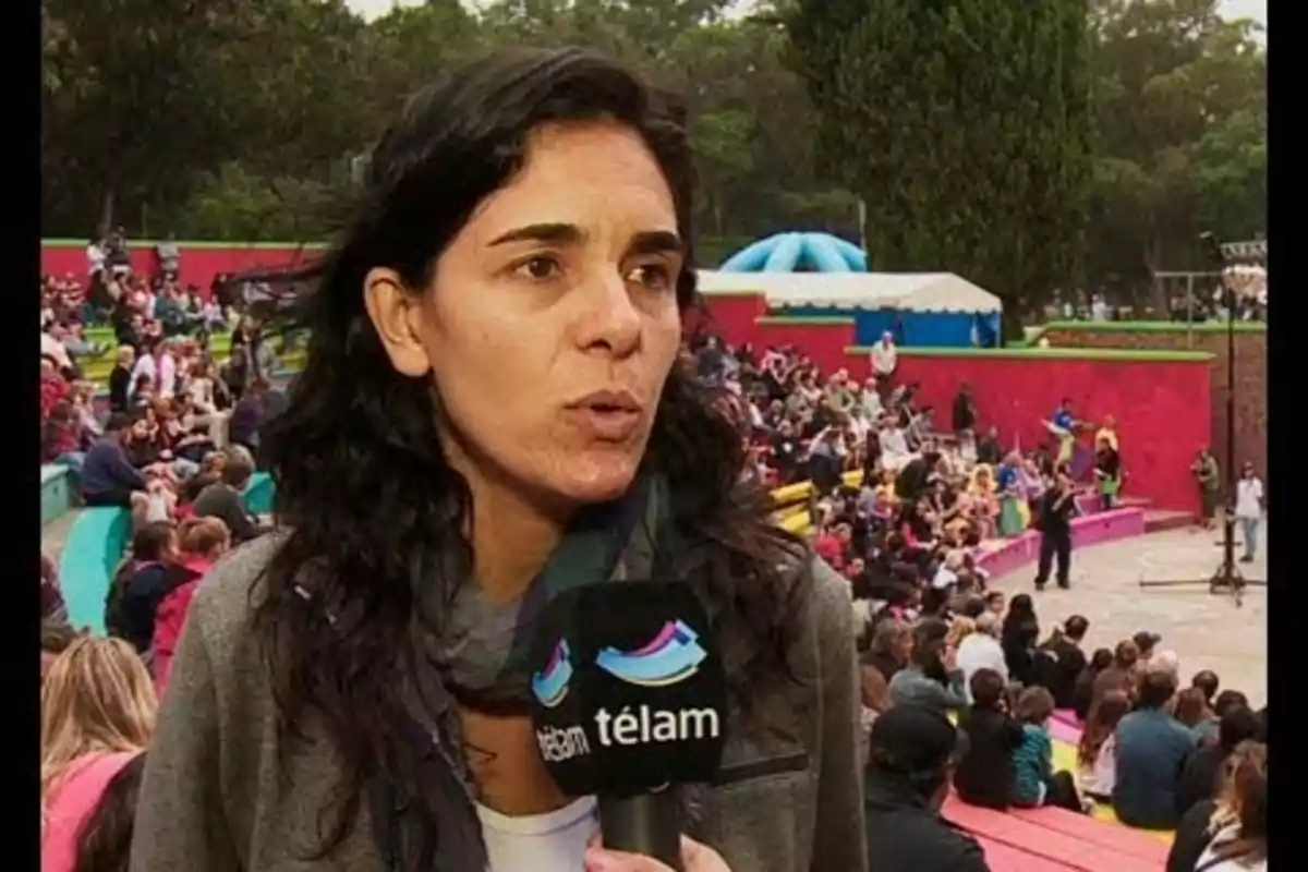 Una mujer siendo entrevistada en un evento al aire libre con un micrófono de Télam, rodeada de un público sentado en gradas coloridas.