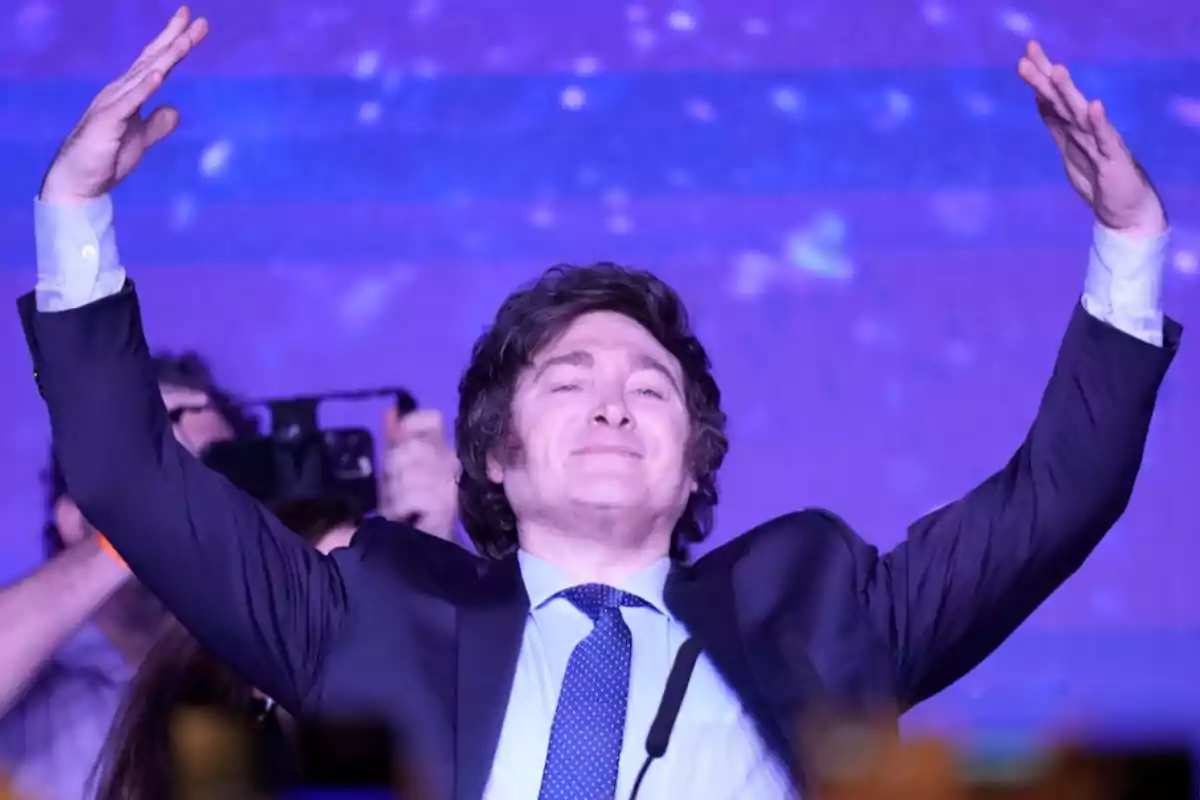 Un hombre con traje y corbata levanta los brazos en un escenario con fondo morado. Un hombre con traje y corbata levanta los brazos en un escenario con fondo morado.