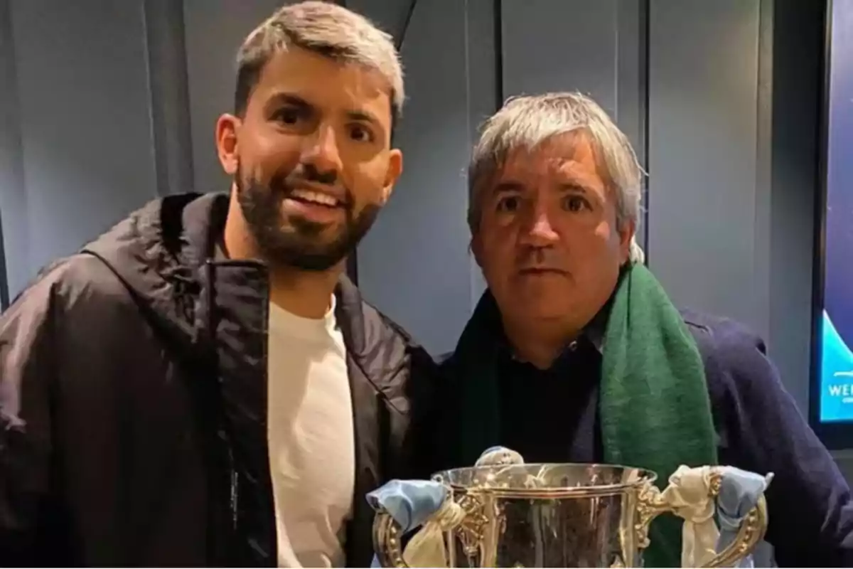 Two people pose together holding a silver trophy in an indoor setting. Two people pose together holding a silver trophy in an indoor setting.